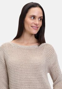 Beige gebreide trui met een brede hals, lange mouwen en een losse pasvorm. Heeft een textuurpatroon en ribbels aan de manchetten.