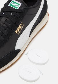 Baskets noires avec un dessus en daim et en textile, logo et bande blanche Puma, semelle en caoutchouc marron, accompagnées de deux lacets blancs.