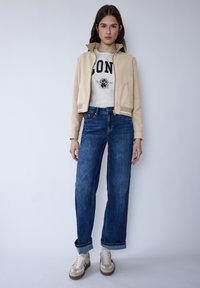 Veste beige courte zippée portée sur un sweat-shirt blanc avec texte noir, associée à un jean bleu taille haute et des baskets crème avec des accents marron.