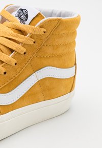 Gul ruskind Vans højt skåret sneaker med hvid stribe, hvid sål og gule snørebånd på hvid baggrund.