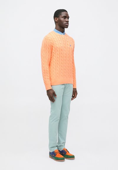 Πορτοκαλί πουλόβερ πλεκτό με σχέδιο καλώδιο, πουκάμισο με ανοιχτό μπλε γιακά, παντελόνι μέντας πράσινο και πολύχρωμα παπούτσια με πορτοκαλί, μπλε και πράσινες λεπτομέρειες.