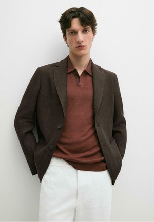 Ung mand med kort mørkt hår iført en brun blazer, rustfarvet polo trøje og hvide bukser, stående mod en ensfarvet hvid baggrund.