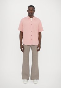 Light pink short-sleeve shirt with wooden buttons, paired with beige flared trousers. Model wearing white sneakers, standing on a white background.