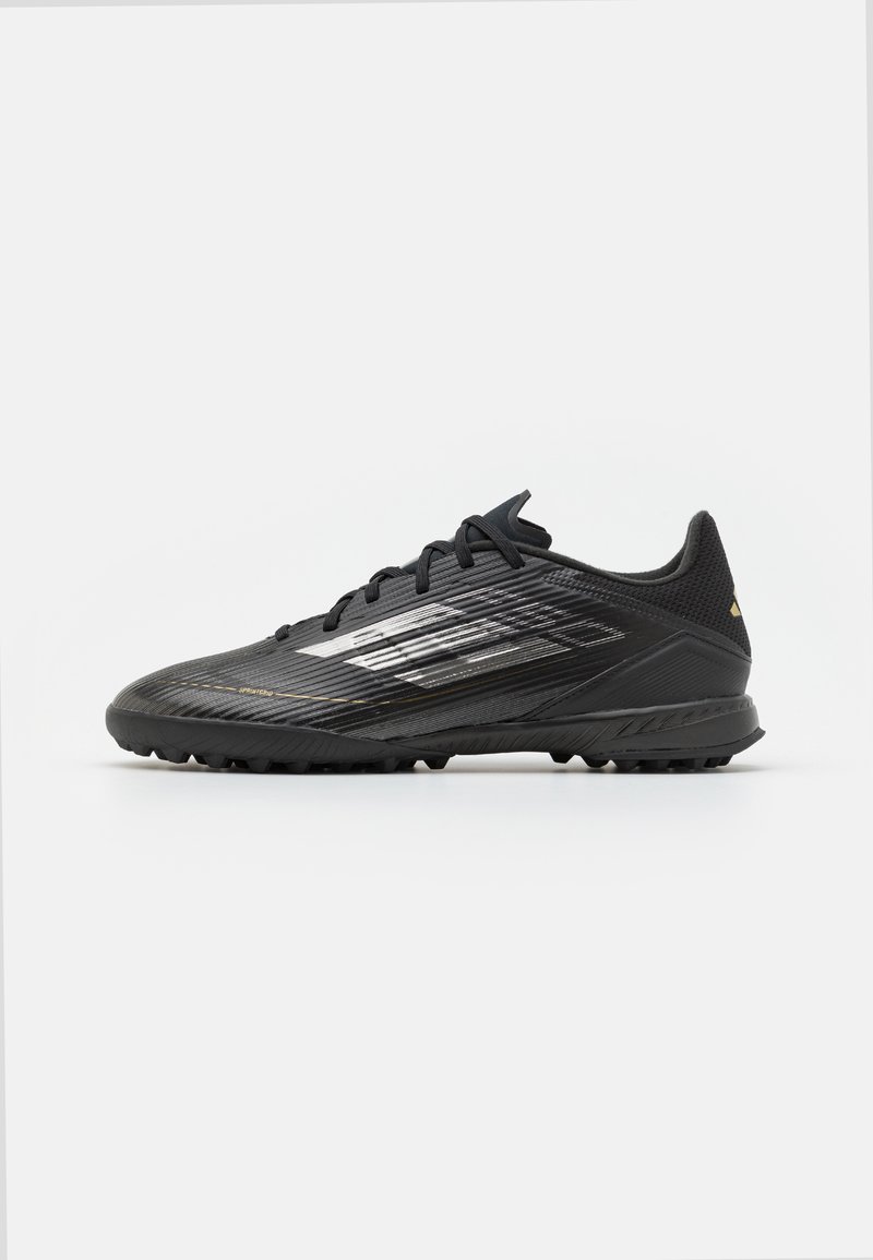 Zapatilla deportiva negra con parte superior texturizada, presentando acentos metálicos y un diseño aerodinámico. Suela con relieve para tracción en superficies de hierba.