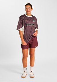 Bordeauxrød sportstrøje med striber, hvide detaljer og logo, kombineret med matchende shorts. Stoffet er åndbart; afslappet pasform for komfort.