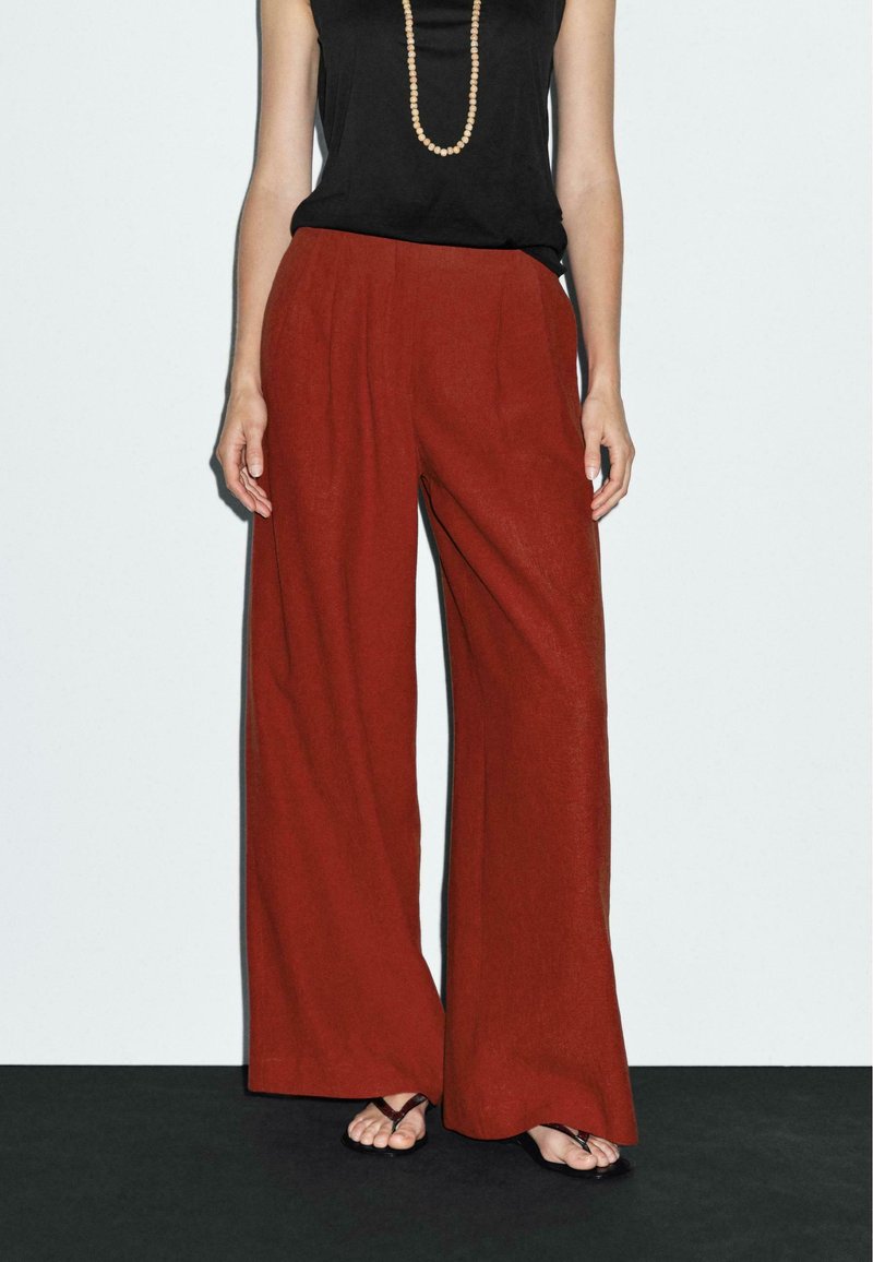 Red wide-leg trousers made of a textured fabric, featuring front pleats, paired with a black top and flat sandals.