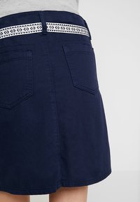 Navy blue skirt with two back pockets and a black-and-white patterned woven belt at the waist, worn with a light gray top.