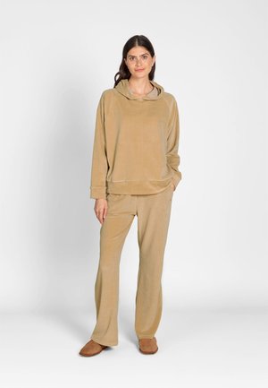 Woman standing wearing beige hooded sweatshirt and matching loose pants with brown slip-on shoes against plain light background.