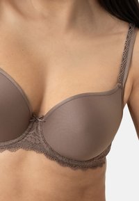 Muotoinen rintaliivit pehmeässä, hillityssä ruskeassa kankaassa. Hieman koristeellinen pitsireunus vyön ja hihnojen ympärillä. Sujuva rakenne, jossa on hieno kiilto.