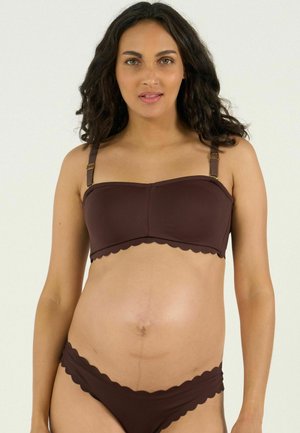 Femme enceinte aux cheveux bouclés et foncés portant un haut bandeau marron et un bas de bikini assorti à bords festonnés sur un fond uni.