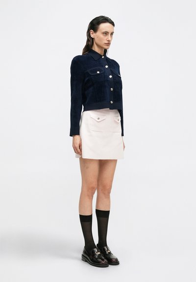 Mujer de pie con una chaqueta azul marino abotonada, falda rosa pálido con bolsillos frontales, medias negras transparentes hasta la rodilla y mocasines negros pulidos.