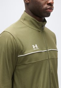 Homme portant une veste zippée Under Armour vert olive avec un détail de piping blanc à travers la poitrine, visible de l'épaule au menton.
