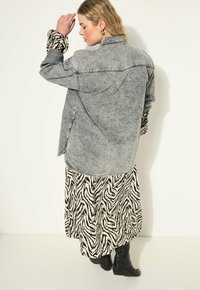 Šedá denimová oversized košile s límečkem, vrstvená přes dlouhé krémovo-černé šaty se zebřím vzorem, doplněná nadkolenkovými černými botami.