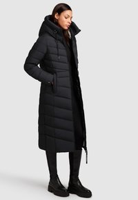 Lungo piumino nero con cappuccio, caratterizzato da cuciture verticali, chiusura con zip e tasche laterali. Tessuto liscio con una finitura leggermente lucida.