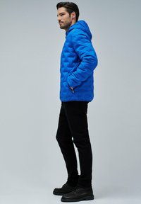Hombre con chaqueta acolchada azul con capucha, pantalones negros y zapatos negros de pie de perfil con las manos en los bolsillos contra un fondo liso.