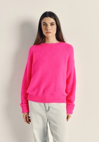 Sieviete ar gariem tumšiem matiem, valkājot spilgti rozā džemperi un gaiši zilas džinsa bikses, stāv pret vienkāršu smilškrāsas fonu.