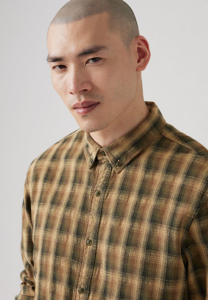 Camisa de cuadro marrón y verde con un tejido texturizado, que cuenta con un collar y botones visibles en la parte delantera.