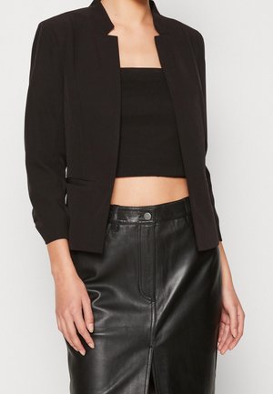 Kvinde iført sort kort blazer, sort tube top og sort lædernederdel med høj talje, frontslids og knaplukning.