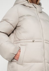 Chaqueta acolchada gris claro con capucha, cremallera en el frente y bolsillos laterales. Textura suave con detalles de costura a lo largo de las costuras.