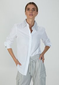 Weißes Hemd mit strukturiertem silbernen Kragen, langen, bis zu den Ellenbogen gerollten Ärmeln und schwarzen Knöpfen an den Manschetten. Glattes Material.