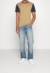 Man som bär en beige och svart kortärmad t-shirt, ljusblå denimjeans och vita sneakers, stående med ena handen i fickan.