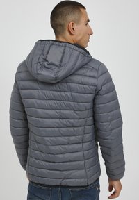 Siva puhovka s kapuco, ki ima vodoravne prešite odseke, gladko teksturo in elastične manšete, oblečena čez jeans.