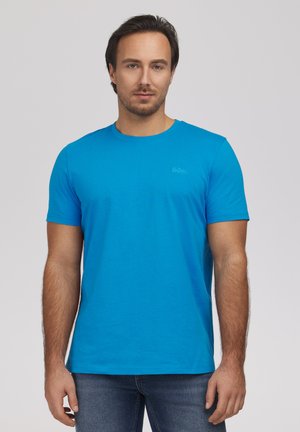 Mężczyzna z krótkimi ciemnymi włosami, ubrany w jednolity, jaskrawoniebieski, krótkim rękawem T-shirt i niebieskie dżinsy, stojący na jednolitym jasnym tle.