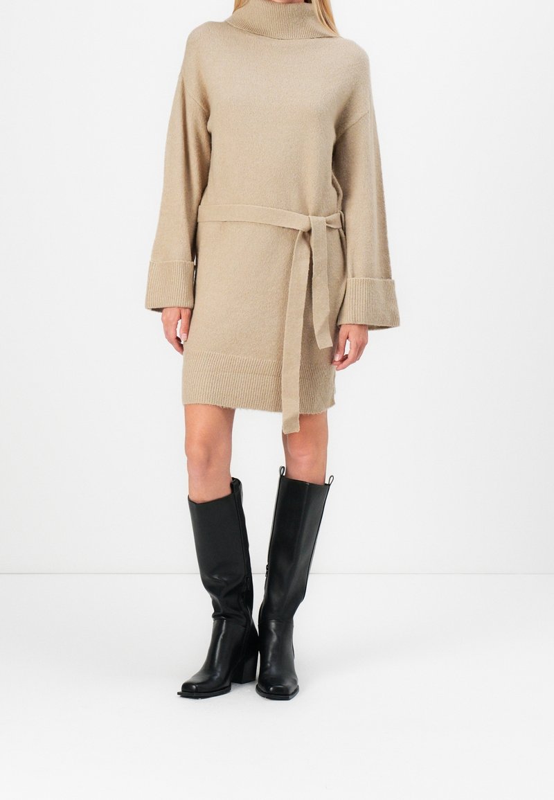 Robe-pull beige en maille avec un col montant et une taille ceinturée. Manches longues avec poignets côtelés. Assorti à des bottes en cuir noires montantes jusqu’au genou.