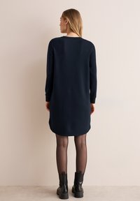 Granatowa sukienka z długim rękawem, z falbaniastym dołem i okrągłym dekoltem, zestawiona z czarnymi, masywnymi botkami. Gładka tekstura materiału.