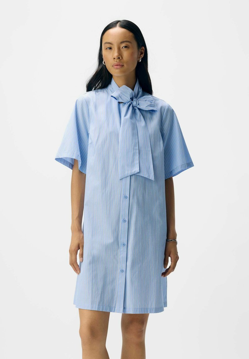 Abito camicia a righe blu chiaro con fiocco al colletto, maniche corte e chiusura con bottoni. Tessuto morbido con una vestibilità comoda.