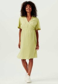 Vestido envolvente verde claro de manga corta con un acabado texturizado, cintura ceñida y falda que cae hasta media pantorrilla. Zapatillas blancas completan el look.