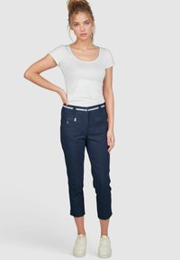 Marineblaue, taillierte Cropped-Hosen mit zwei Fronttaschen, silbernen Hardware-Akzenten und einem weißen gestreiften Bund. Kombiniert mit einem weißen T-Shirt.