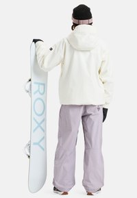 Snowboardoutfit bestaande uit een crème jas en lavendelkleurige broek, terwijl hij een witte snowboard vasthoudt met "ROXY" in lichtblauwe letters.
