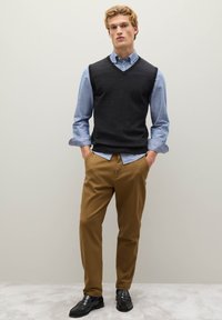 Grauer ärmelloser Pullover über einem hellblauen Hemd mit Knöpfen, beigefarbene Hose und schwarze Schuhe; das Modell steht vor einem neutralen Hintergrund.