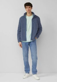 Fleecejack met een hoge kraag in zacht blauw, met een rits aan de voorkant en twee zijzakken, gedragen over een licht mintgroen t-shirt en gecombineerd met licht denim jeans.