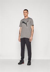 T-shirt de sport gris avec logo Puma noir, associé à un pantalon sombre et des baskets noires avec des accents rouges. Le mannequin se tient avec les bras détendus.