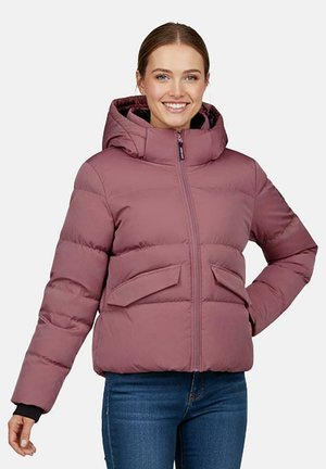Jonge vrouw lacht en draagt een mauve gewatteerd jack met capuchon en een blauwe spijkerbroek, staand tegen een effen lichte achtergrond.