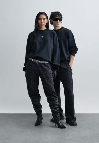 Melnas, lielizmēra džemperi ar logotipu, kombinēti ar rakstainiem melniem biksēm. Aksesuāri ietver sudraba jostu un melnus zābakus līdz ceļiem.