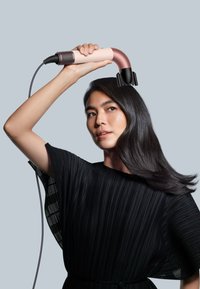 Hårfön med en böjd design, rosa och svarta färger, texturerat grepp och munstycke. Används av en person med långt, rakt hår.