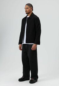 Zwarte jas met knoopsluiting, wit T-shirt eronder, losse zwarte jeans en zwarte schoenen met gekleurde veters. Eenvoudig, casual ontwerp.