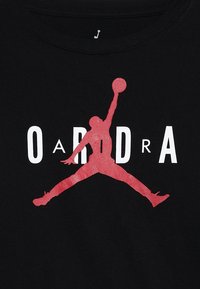 Schwarzes Baumwoll-T-Shirt mit einer roten Silhouette einer springenden Figur und weißem Text "O a R D R A", der auf der Brust zentriert ist.