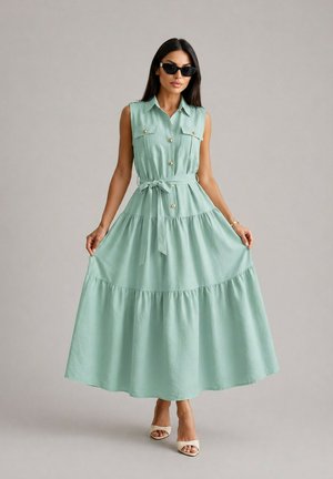 Femme portant une robe sans manches vert clair à boutons avec ceinture et lunettes de soleil, tenant les côtés de la jupe, debout sur un fond gris.