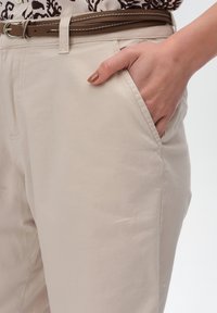 Beżowe spodnie z bawełny z bocznymi kieszeniami, płaskim przodem i brązowym paskiem z białym ściegiem, prezentujące gładką fakturę.