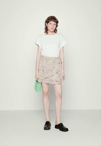 T-shirt branca, saia envolvente lilás com padrão floral, sapatos pretos tipo slip-on e uma pequena bolsa verde. Tecido suave, volantes em camadas, design casual.