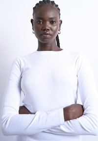 Νεαρή μαύρη γυναίκα με πλεκτά μαλλιά και σκουλαρίκια-κρίκους, φορώντας λευκό πουκάμισο με μακριά μανίκια, με σταυρωμένα χέρια και ουδέτερη έκφραση, λευκό φόντο.