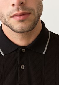 Zwart poloshirt met een gestructureerd patroon, voorzien van een standaardkraag en een knoopsluiting met drie knopen, geaccentueerd door grijze strepen op de kraag.