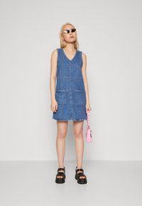 Denim haljina u plavoj boji s V-izrezom, dva prednja džepa i sirovim rubom. Uparena s masivnim crnim sandalama i malom ružičastom torbicom.
