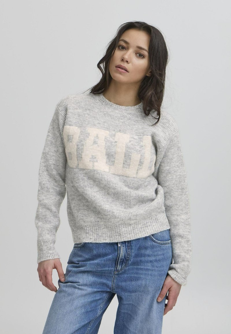 Grå strikkegenser med ribbestruktur, med en stor kremfarget "BAD" grafikk på fronten. Kombinert med blå denimjeans.