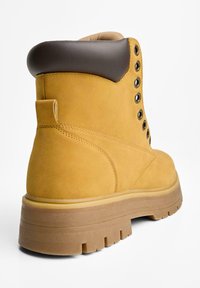Gelbe Wildleder-Stiefeletten mit einem braunen gepolsterten Kragen, metallischen Ösen und einer dicken Gummisohle mit tiefen Profilrillen.