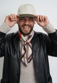 Beige Kappe mit olivgrünem Schirm, schwarze Lederjacke, grauer Strickpullover und gemusterter Seidenschal mit Streifen in Braun und Orange.